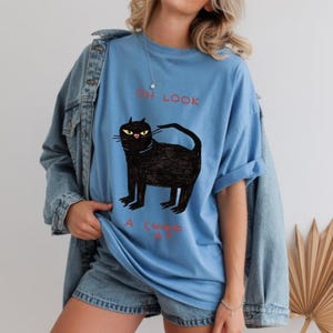 Pu&ograve; includere: T-shirt azzurra con un'illustrazione di un gatto nero e la scritta "OH LOOK A CAT". Il gatto ha gli occhi gialli e un design stravagante. La maglietta &egrave; realizzata in tessuto morbido.