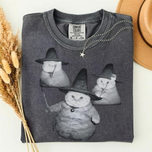 May include: Dark gray t-shirt featuring a graphic of three cats wearing witch hats. The cats are depicted in a grayscale image. A silver necklace with a star pendant is draped on the shirt. The shirt has a Comfort Colors tag.