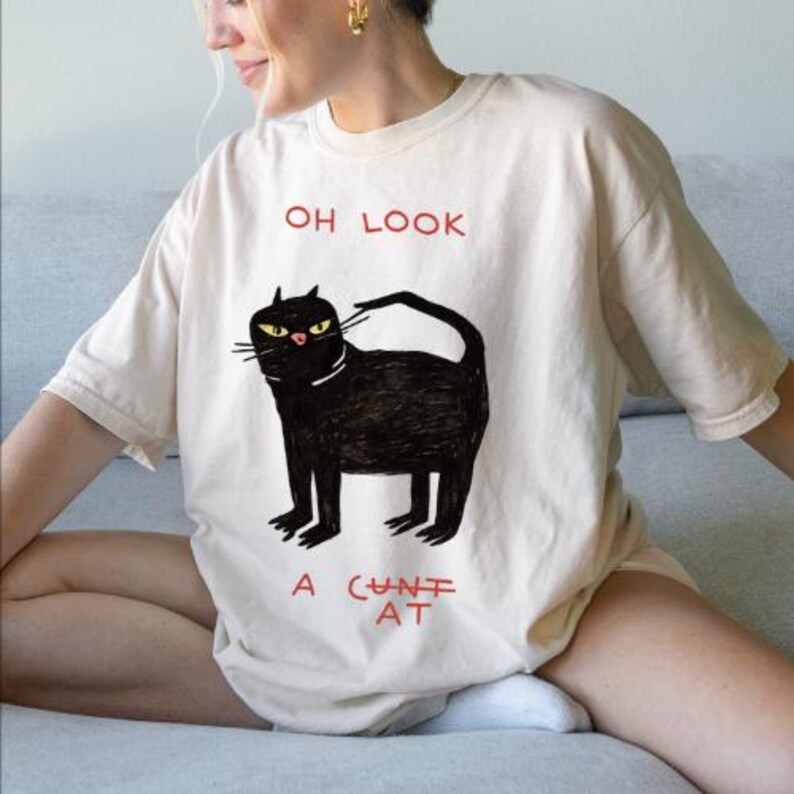 Pu&ograve; includere: T-shirt color crema con illustrazione di un gatto nero con occhi gialli e la scritta rossa "OH LOOK" sopra e "A CUNT AT" sotto. Il gatto ha un collare bianco.