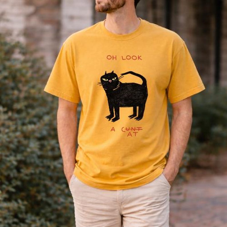 Pu&ograve; includere: T-shirt giallo senape con grafica di un gatto nero. Il testo "OH LOOK" &egrave; sopra il gatto, e "A CUNT AT" sotto. Il gatto ha una coda ricurva.