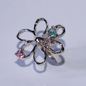 Puede incluir: Anillo plateado con forma de flor de cinco pétalos. Cada pétalo está adornado con pequeñas piedras preciosas rosas, azules y moradas. Las piedras están engastadas en los pétalos plateados, creando un diseño floral brillante.