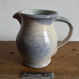 以下が含まれることがあります： 丸みを帯びたボディ、注ぎ口、湾曲したハンドルを備えた、まだらのセラミックピッチャー。ピッチャーは、青い斑点と青い縁が付いた明るいベージュのベースが特徴です。ハンドルと注ぎ口は明るい青緑色です。ピッチャーは木製の表面に置かれています。