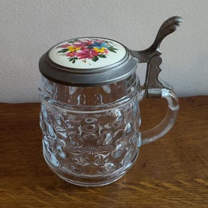 May include: A clear glass beer stein with a floral decorated lid. The lid has a white background with a colourful arrangement of pink, yellow, and blue flowers. It has a metal hinged lid and handle.