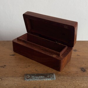 May include: Open, rectangular wooden box with a hinged lid. The box is made of dark brown wood and has a smooth, polished finish. The interior features a small, raised compartment. The box is sitting on a wooden surface.