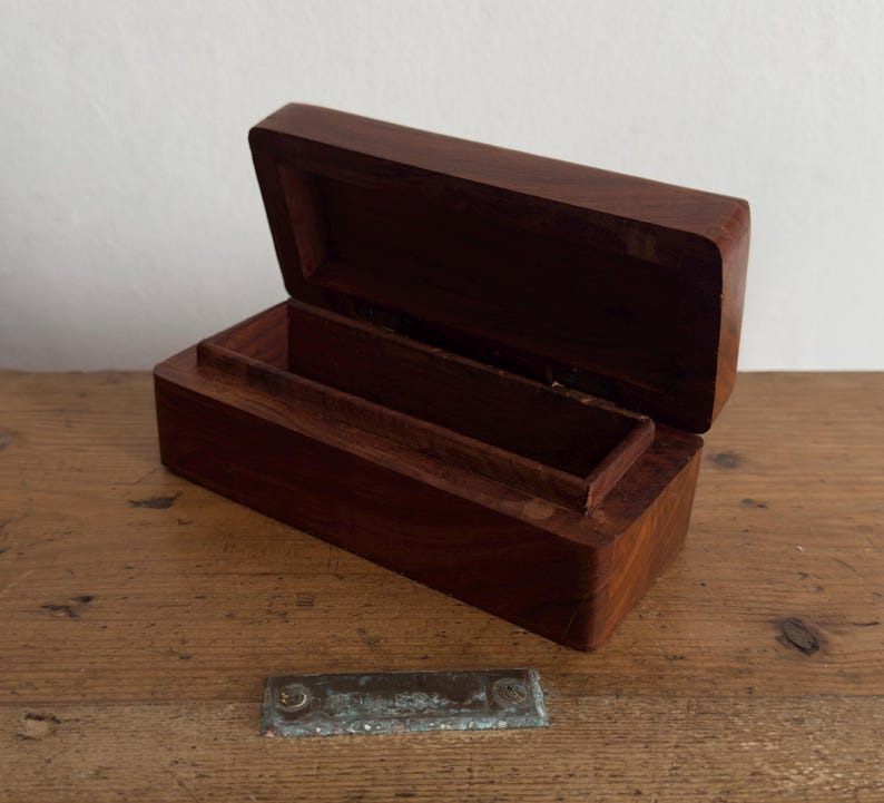 May include: Open, rectangular wooden box with a hinged lid. The box is made of dark brown wood and has a smooth, polished finish. The interior features a small, raised compartment. The box is sitting on a wooden surface.