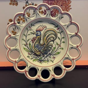 May include: A decorative ceramic plate with a rooster design. The plate has a scalloped edge with circular cutouts and a hand-painted rooster in the centre, surrounded by floral motifs. The plate's rim is accented with a pink border.