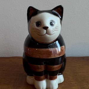 May include: A ceramic cat figurine with a white face, brown nose, and black ears. The cat's body is black with brown stripes. The figurine has white paws and is sitting upright. The cat has a smiling expression.
