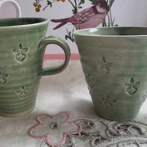 以下が含まれることがあります： 緑色の花柄のセラミックマグカップ2つ。 マグカップは、幅広でわずかにフレアしたリムと湾曲したハンドルを備えています。 花柄は表面にエンボス加工されています。 マグカップは、花柄のある明るい色の表面にあります。