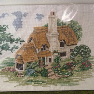 May include: Cross-stitch picture of a cottage with a thatched roof, chimney, and surrounding greenery. The artwork features a detailed design with various shades of green, brown, and beige, creating a picturesque scene. The cottage is framed and ready to hang.