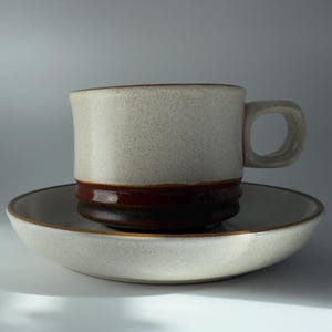 以下が含まれることがあります： 濃い茶色のバンドとハンドルが付いた、斑点のあるベージュ色のセラミックマグが、お揃いのソーサーの上に置かれています。マグとソーサーは、コーヒーまたは紅茶に適した、シンプルでクラシックなデザインです。セットはセラミック製です。