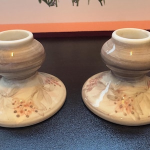 May include: A pair of ceramic candle holders. Each has a rounded top and a wide, oval base. The tops are brown with a white rim, and the bases are cream-coloured with a floral design in pink and brown.