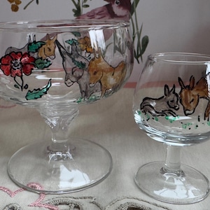 May include: Two clear glass goblets with hand-painted donkey and floral designs. The larger goblet features a red flower and various donkeys in shades of brown and grey. The smaller goblet has two donkeys. Both have a stem and a round base.