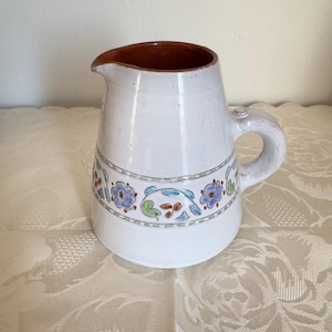 May include: A white ceramic jug with a terracotta-coloured interior and a side handle. The exterior features a hand-painted horizontal band decorated with blue flowers, green leaves, and brown accents, set between two thin green lines.
