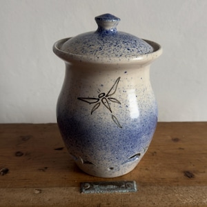 May include: A ceramic jar with a lid, in a blue and white speckled design. A black dragonfly is painted on the front. The jar has a rounded shape with small holes near the base. The lid has a small knob on top.