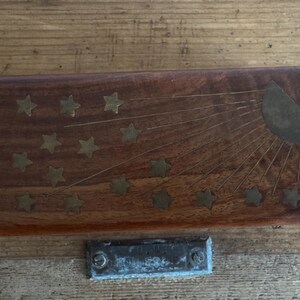 May include: A rectangular wooden box with a sunburst and star design. The box is made of dark wood and has brass inlays forming a sunburst and star pattern. A small, metal plate is below the box.