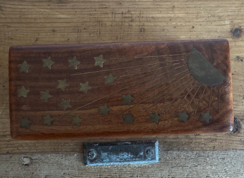 May include: A rectangular wooden box with a sunburst and star design. The box is made of dark wood and has brass inlays forming a sunburst and star pattern. A small, metal plate is below the box.