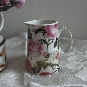 May include: A white ceramic mug with a black handle and the text "But First, Coffee" in a black, handwritten-style font. The mug is on a white surface.