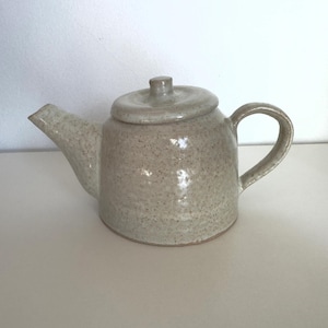 May include: A ceramic teapot with a speckled light beige glaze. The teapot features a rounded body, a curved handle, a short spout, and a matching circular lid with a small knob handle on top. The surface has a glossy, textured finish.
