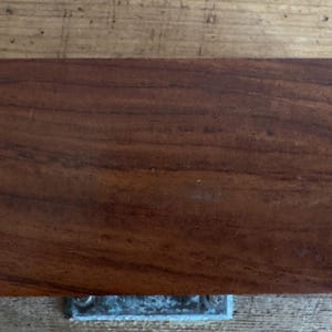 May include: A rectangular, solid wood object with a rich, dark brown color and visible wood grain. The object has rounded corners and a metal fixture attached to the bottom. The wood appears to be polished, highlighting the natural patterns.