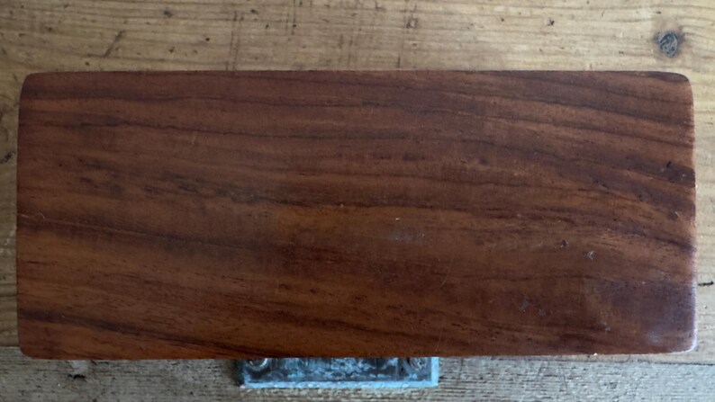 May include: A rectangular, solid wood object with a rich, dark brown color and visible wood grain. The object has rounded corners and a metal fixture attached to the bottom. The wood appears to be polished, highlighting the natural patterns.