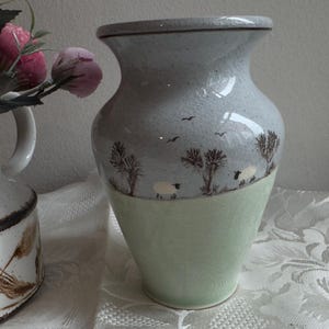 May include: A ceramic vase with a pale green base and a grey upper section. The grey section features a landscape design with trees, sheep, and birds. The vase has a flared top and a textured surface.