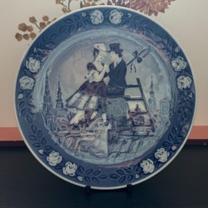 May include: A blue and white porcelain plate with a romantic scene of two figures and a city backdrop. The rim is decorated with white roses and foliage. A classic decorative plate, suitable for display or collection.