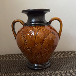 May include: A ceramic vase with a brown glaze and a black rim and base. The vase features two handles and embossed floral designs. The vase is sitting on a patterned surface.