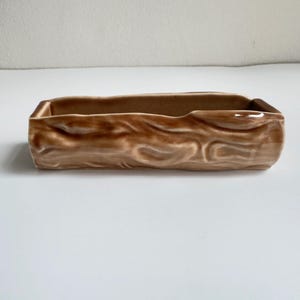 May include: A rectangular ceramic planter with a glossy brown finish and a textured exterior that mimics the appearance of natural tree bark. The vessel features an open top and a smooth interior, designed with a long, narrow, horizontal shape.