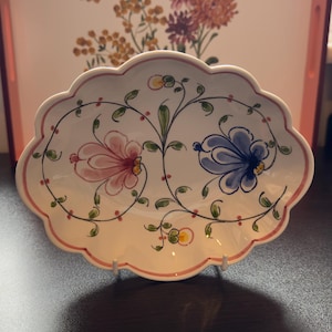 May include: A white, scalloped-edge ceramic serving dish with a pink and blue floral design. The dish has a pink rim and is supported by a white stand. The floral pattern includes pink and blue flowers with green leaves and vines.