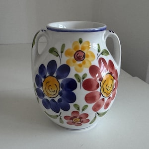 May include: White ceramic vase with two handles, decorated with hand-painted flowers in blue, yellow, and red. Green leaves complement the floral design. The vase has a blue rim and a rounded shape, ideal for home decor, measuring approximately 20 cm in height.