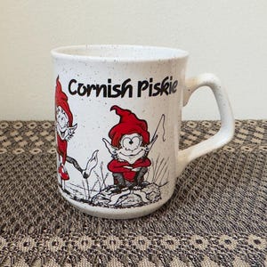 May include: White ceramic mug with a speckled finish, featuring the text "Cornish Piskie" in black. The mug showcases two cartoon figures in red outfits and hats. The mug has a curved handle and is set on a patterned surface.
