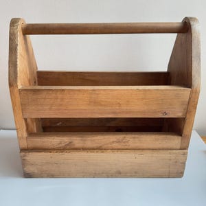 May include: A rectangular wooden caddy featuring a natural light brown wood grain finish. The structure includes a solid base, two horizontal side slats, and a sturdy rounded handle spanning the top between two upright vertical end panels.