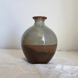 Peut inclure: Un petit vase en céramique avec un corps arrondi et un col étroit. La partie supérieure présente un vernis gris clair brillant, tandis que la partie inférieure est finie dans un brun mat profond. Les deux couleurs se rejoignent en une ligne.