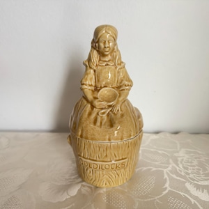 May include: A tan ceramic figurine of a person with long hair, wearing a dress and holding a small bowl and spoon. The figure sits atop a cylindrical base with a wood-grain texture, featuring the embossed text GOLDILOCKS on the front side.
