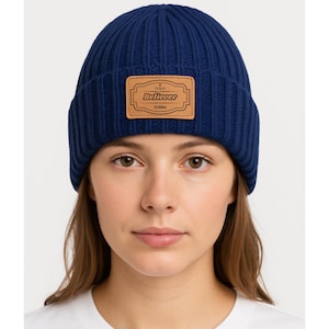 May include: A navy blue ribbed knit beanie hat with a rectangular leather patch. The patch features the word "Believer" in a decorative font, with the name "Debbie" below. A classic winter hat.