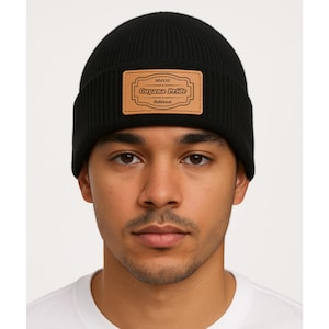 May include: A black beanie with a rectangular leather patch. The patch features the text "MMXXV Guyana Pride Robinson" in a decorative font. The beanie is made of a ribbed knit material.