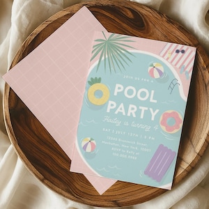 Pode incluir: Um convite para uma festa na piscina com um fundo azul claro, apresentando um gráfico de piscina com um flutuador de abacaxi, um flutuador de donut e bolas de praia. O convite está em uma tigela de madeira com um envelope rosa.