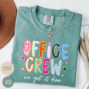 May include: Light green Comfort Colors t-shirt with the words "OFFICE CREW" in colorful, glitter-outlined letters. The shirt also features a smiling face, stars, and the phrase "we get it done". A silver necklace with a pendant is draped on the shirt.