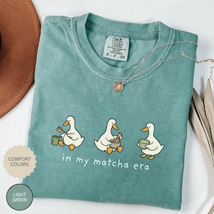 May include: Light green Comfort Colors t-shirt with a graphic of three ducks preparing matcha. The text "in my matcha era" is below the ducks. A silver necklace and a brown hat are also visible.