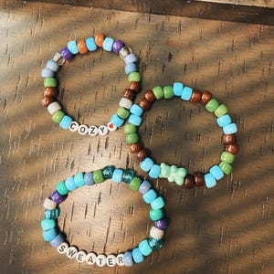 May include: Three colorful beaded bracelets. One bracelet spells "COZY" with white letter beads and a red heart bead. Another bracelet spells "SWEATER". The bracelets feature various colors including blue, green, brown, and purple.