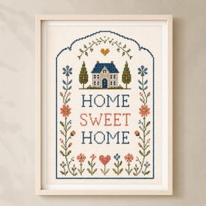 May include: A framed cross-stitch embroidery featuring a house, a small heart, and the text "HOME SWEET HOME" in blue and red thread. The design is bordered by a blue arched frame with floral vines, green trees, and colorful flowers on a cream fabric.