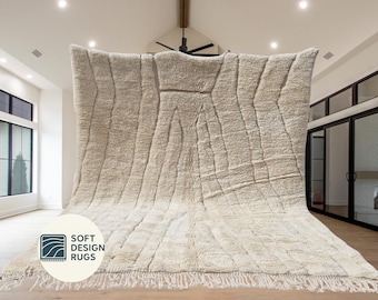 May include: A large, off-white rug with a geometric design and fringe detail. The rug has a textured surface and is displayed in a well-lit room. The rug has the text "SOFT DESIGN RUGS" on it.