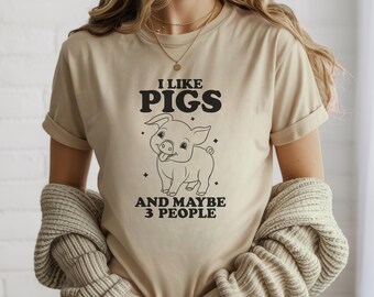 Könnte beinhalten: Beiges T-Shirt mit dem schwarzen Text "I LIKE PIGS AND MAYBE 3 PEOPLE". Darunter eine Illustration eines lächelnden Schweins mit herausgestreckter Zunge. Das Shirt hat umgekrempelte Ärmel.