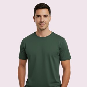 May include: A forest green crew neck t-shirt. The short-sleeved shirt is made of a soft material and is worn by a person. The background is a light pink color.