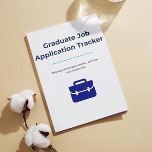 May include: A white notebook titled "Graduate Job Application Tracker" with blue text and a briefcase icon. The notebook is on a beige surface, with cotton sprigs and a patterned glass. The text reads: "Stay organised, apply smarter, and land your dream role."
