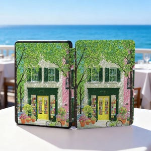 May include: A case featuring a colorful illustration of a bookstore facade with a yellow door, green trim, and potted plants. The design includes a bicycle and a tree with green leaves. The background shows a blue ocean and sky.