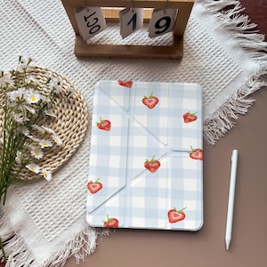 May include: A light blue and white plaid tablet case with a pattern of red strawberries. A white stylus is next to the case. A wooden calendar displays the date, October 19.