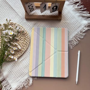 May include: A pastel-striped tablet case in shades of yellow, lilac, green, and pink. A white stylus lies beside the case. A wooden calendar shows "OCT 19". A woven placemat and flowers are also in the image.