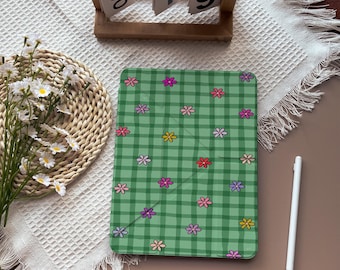 May include: A green tablet case with a checkered pattern and colorful flower designs. The case is on a white surface with a woven placemat and a wooden calendar. A white stylus is also present.
