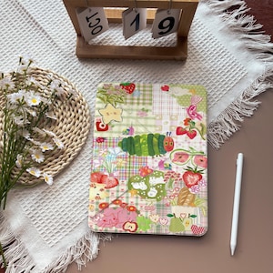 May include: A colorful tablet case with a patchwork design featuring strawberries, a caterpillar, and other whimsical illustrations. The case is on a white surface with a wooden calendar and flowers in the background. A white stylus is also visible.